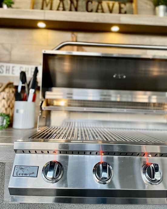 Stainless steel grill with three control knobs on a kitchen counter, with 'Man Cave' sign in the background.